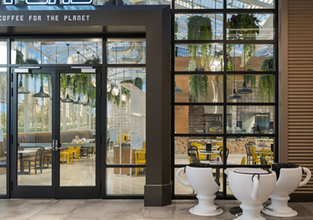 an exterior photo of the glass entry door and front window of a coffee shop with a white table and chairs on the sidewalk